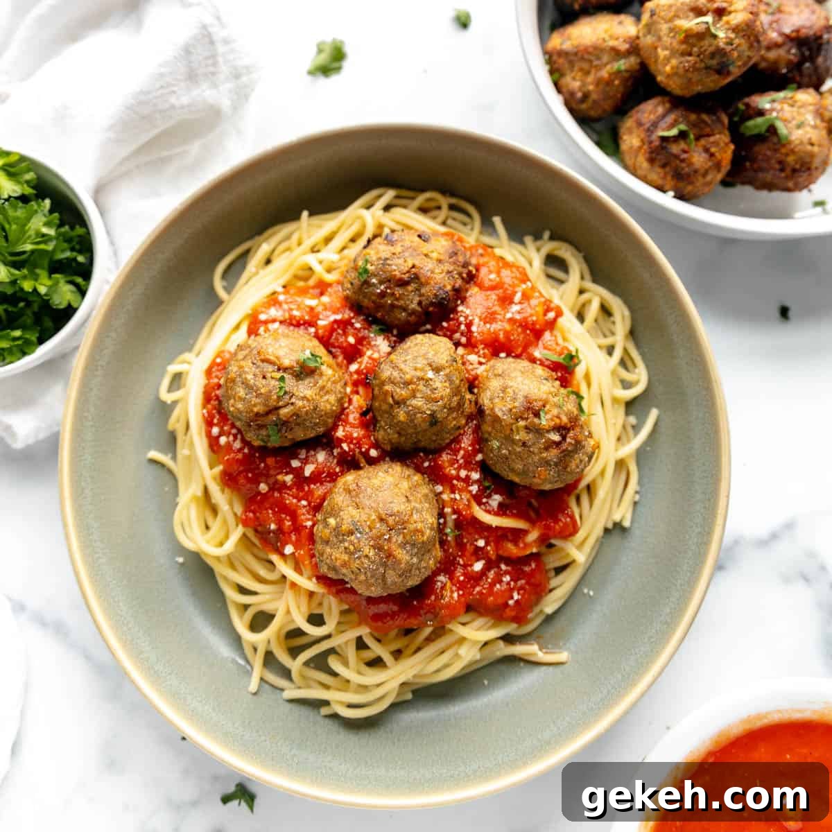 Plate of spaghetti topped with tomato sauce and six meatballs, garnished with herbs. Bowls of extra meatballs, sauce, and parsley are nearby on a white surface.
