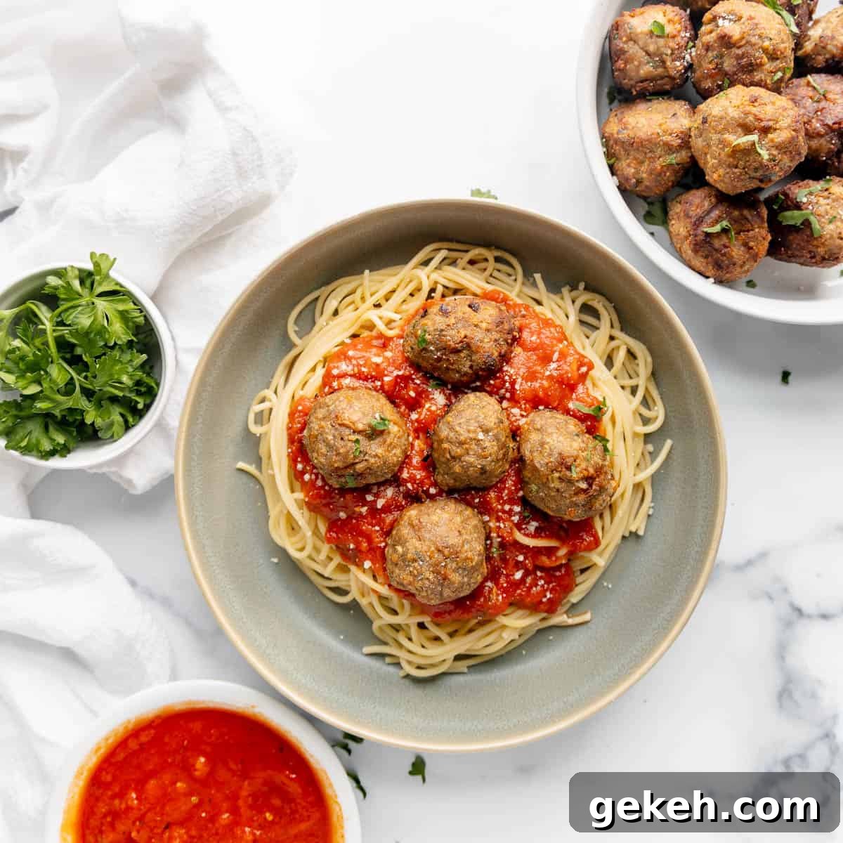 A plate of spaghetti with tomato sauce and meatballs, surrounded by bowls of extra meatballs, fresh parsley, and extra sauce on a marble surface.