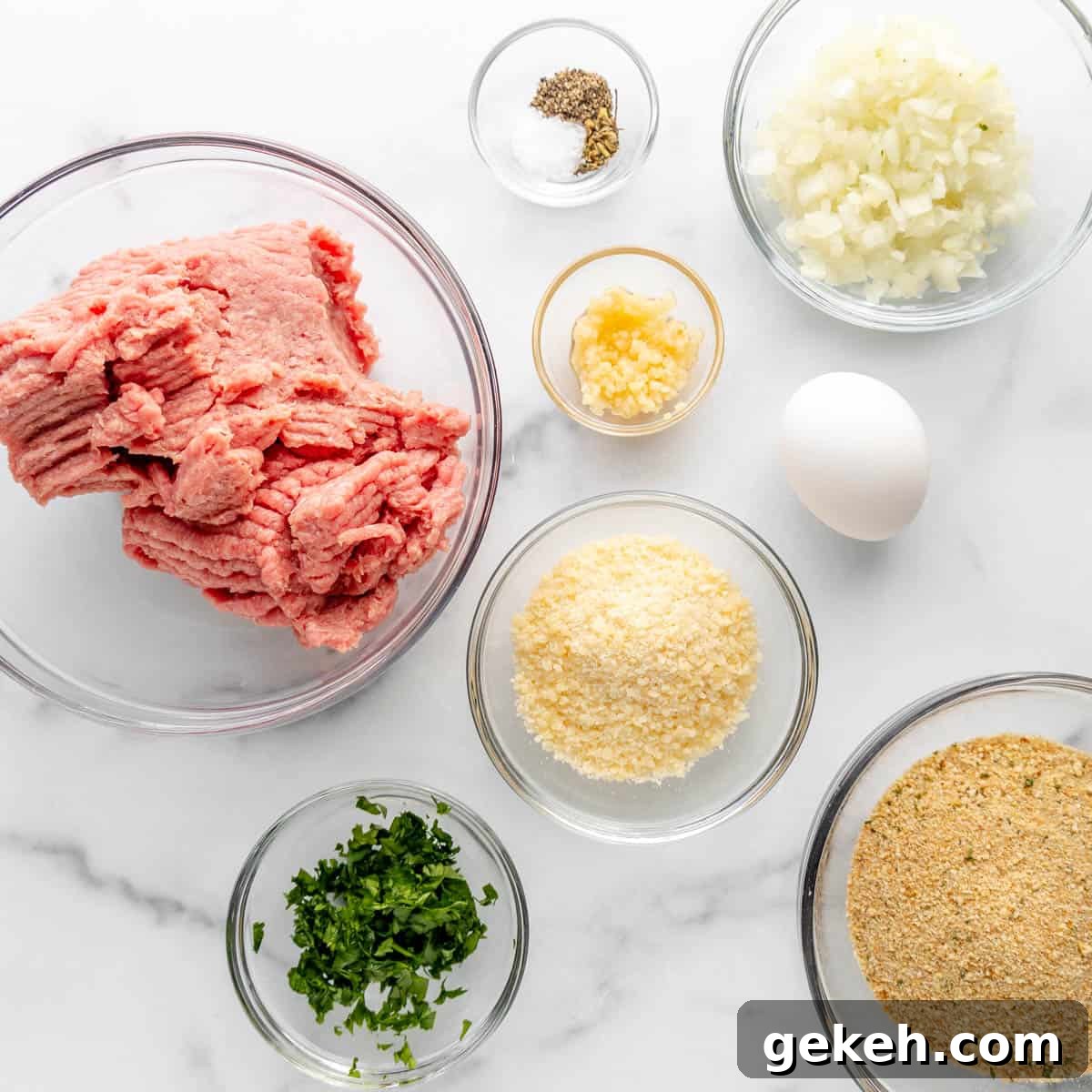Ingredients for a meatball recipe arranged on a marble countertop, including ground meat, chopped onions, garlic, parsley, breadcrumbs, an egg, and seasoning in various bowls.