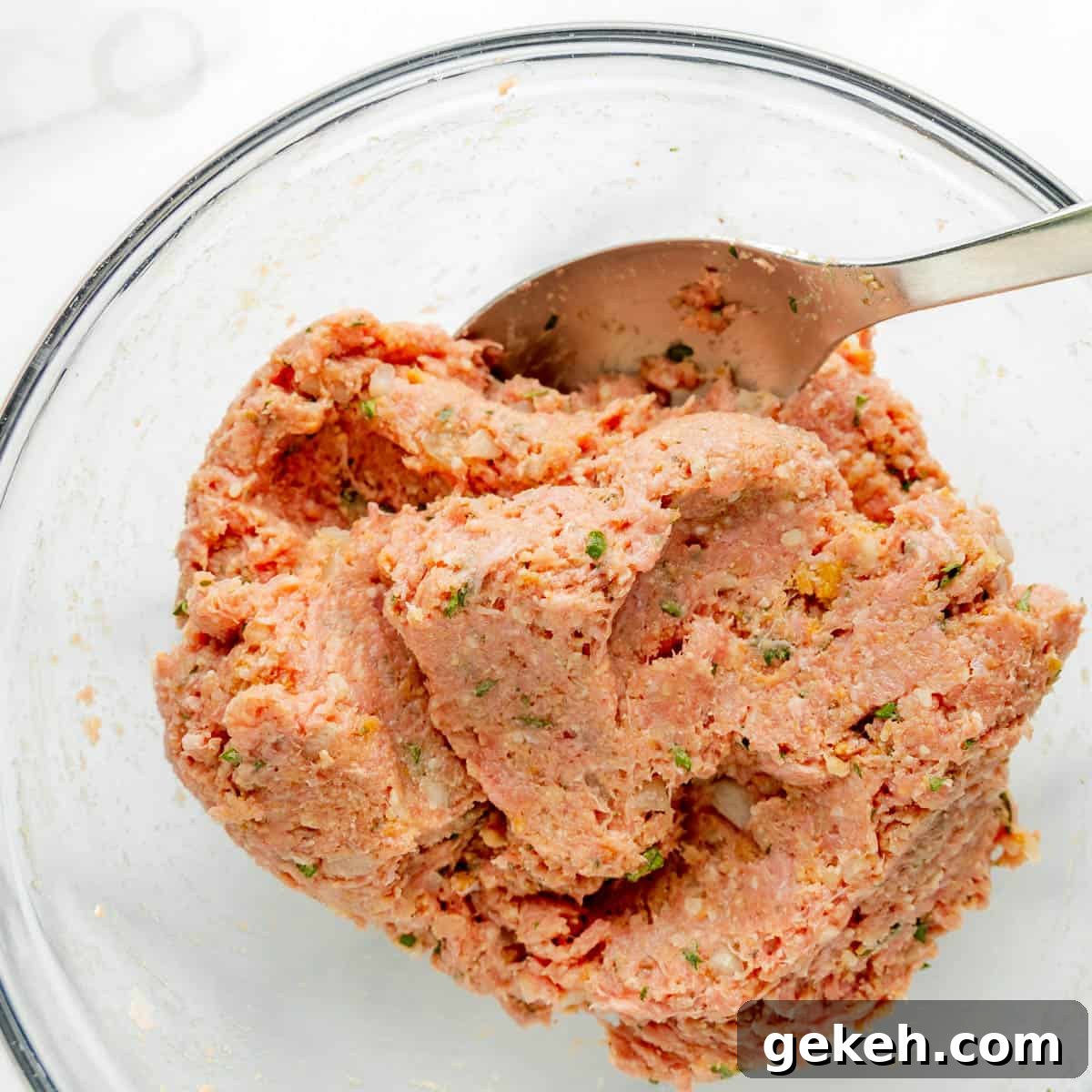 A glass bowl filled with a mixture of ground meat, herbs, and seasonings, with a metal spoon partially submerged in the mixture.