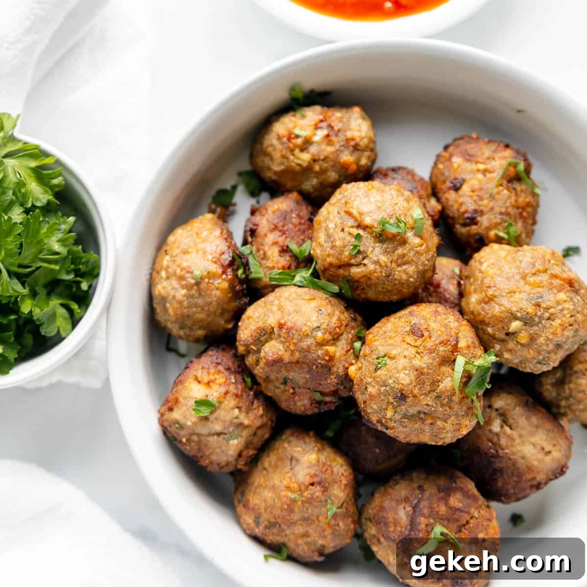 A bowl of golden-brown meatballs garnished with herbs, served next to a small bowl of red sauce and a cup of fresh parsley.