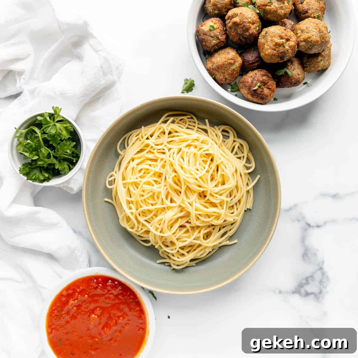 A bowl of cooked spaghetti, a plate of meatballs, a bowl of tomato sauce, and a small bowl of parsley on a marble surface.