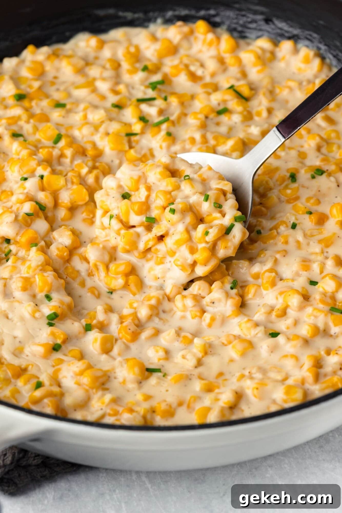 A creamy dish of cheesy corn in a skillet, garnished with chopped chives. A spoon is lifting a portion, showcasing the creamy texture and vibrant yellow corn.