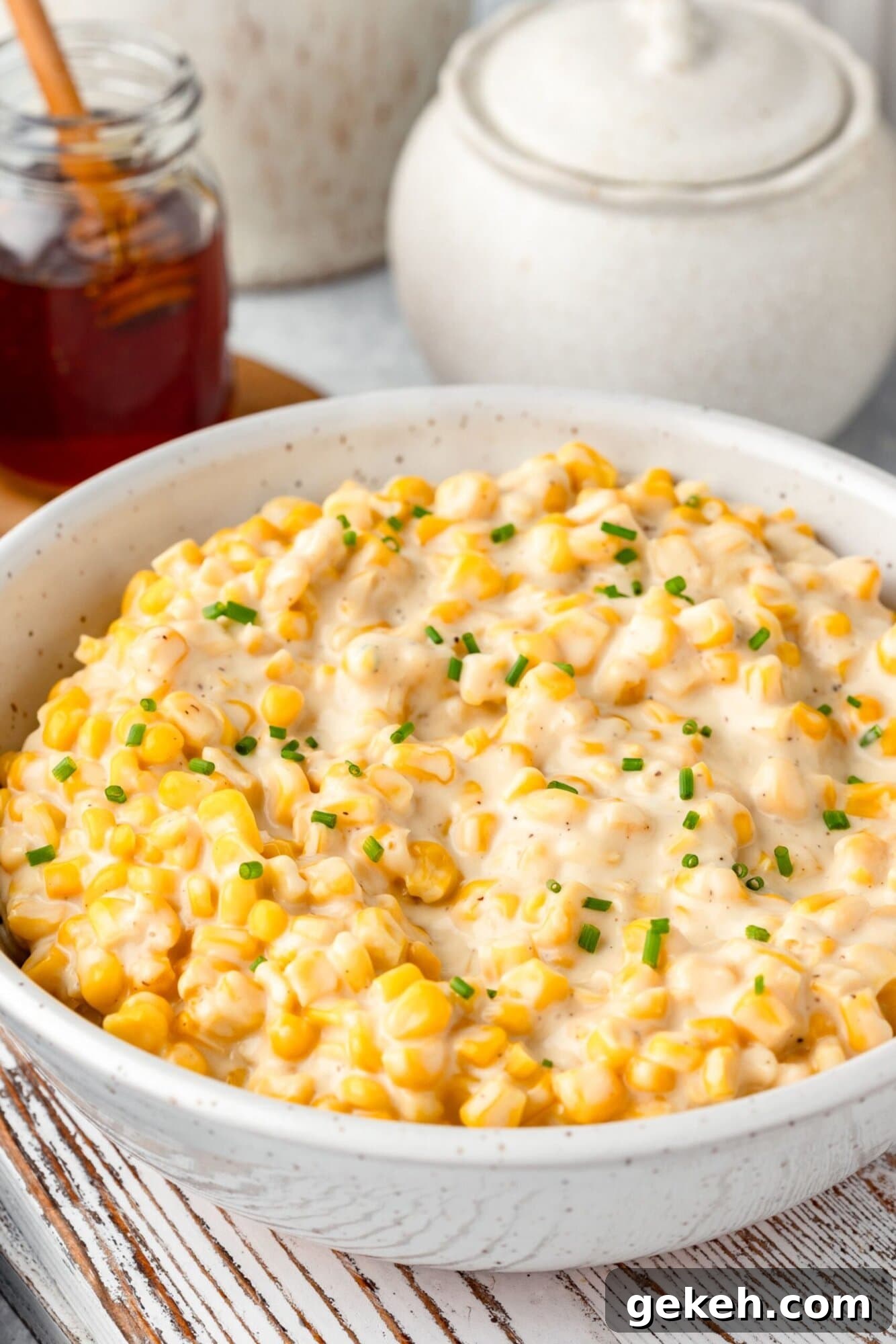 A bowl of creamy corn salad garnished with chopped chives sits on a wooden board. Theres a jar of honey and a ceramic container in the background.
