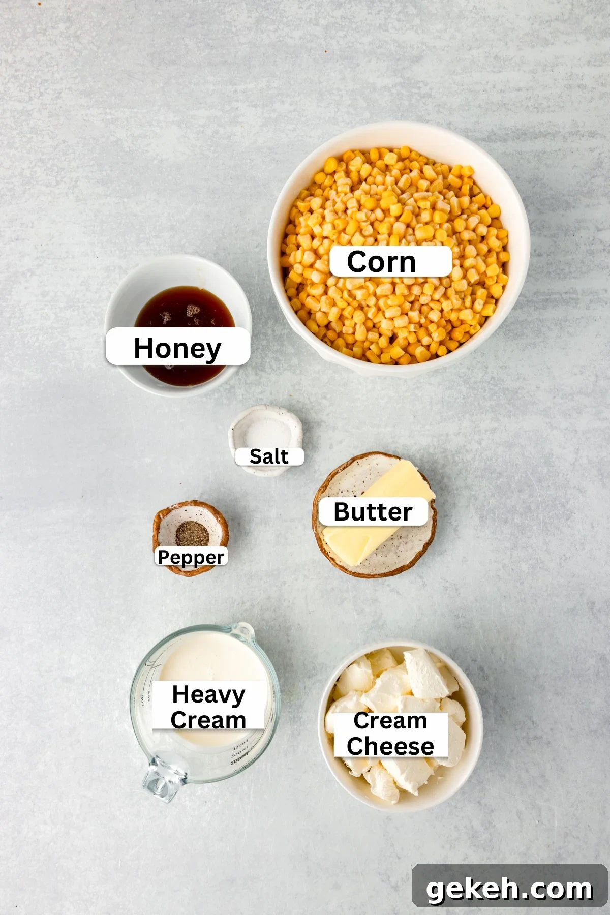 Labeled ingredients on a table include corn in a bowl, honey in a small dish, salt and pepper in separate containers, butter on a slice of wood, heavy cream in a measuring cup, and cream cheese in a bowl.