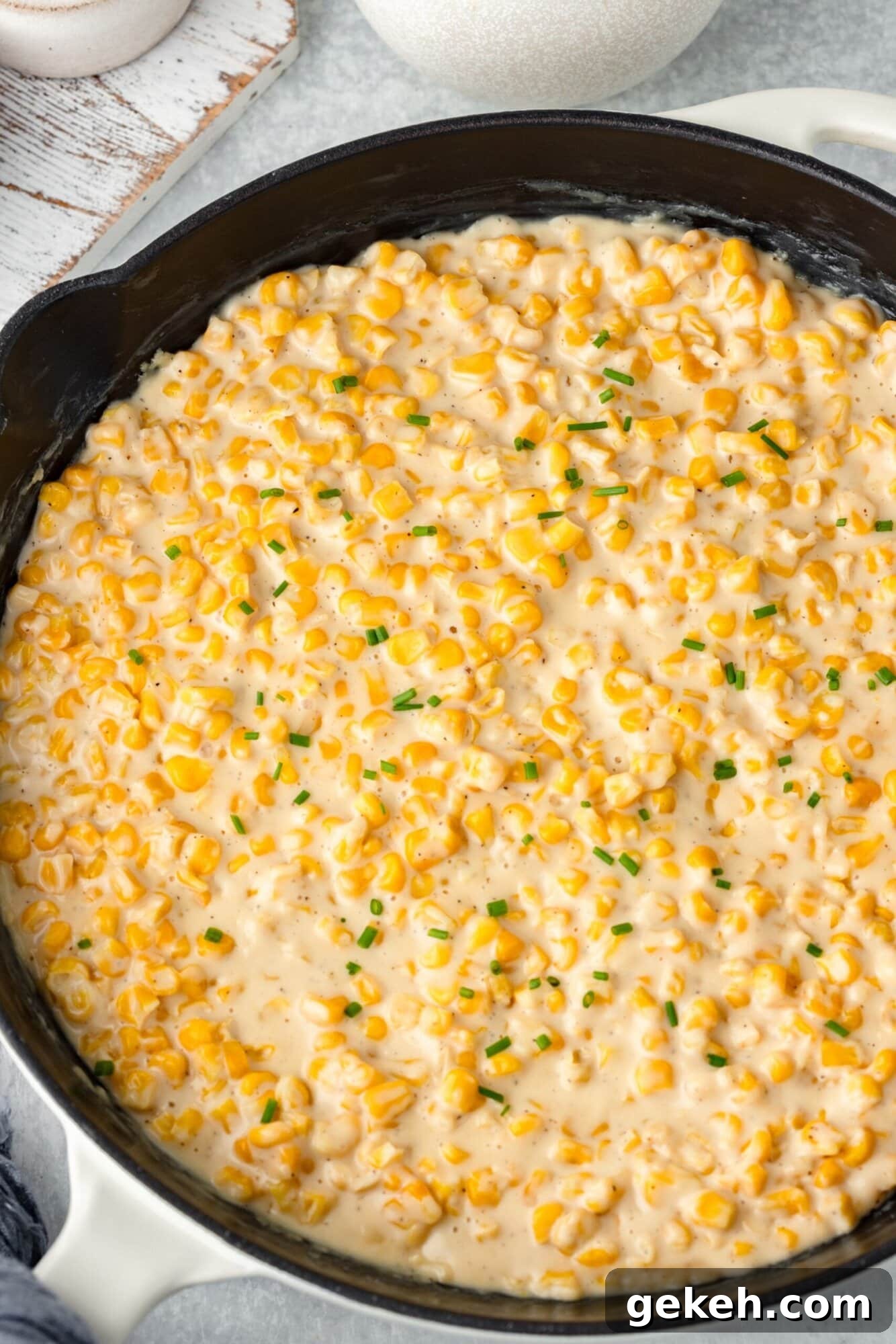 A skillet filled with creamy corn garnished with chopped chives. The dish features a rich, cheesy sauce, showcasing a warm, comforting appearance. The background includes subtle kitchen elements, adding to the cozy ambiance.
