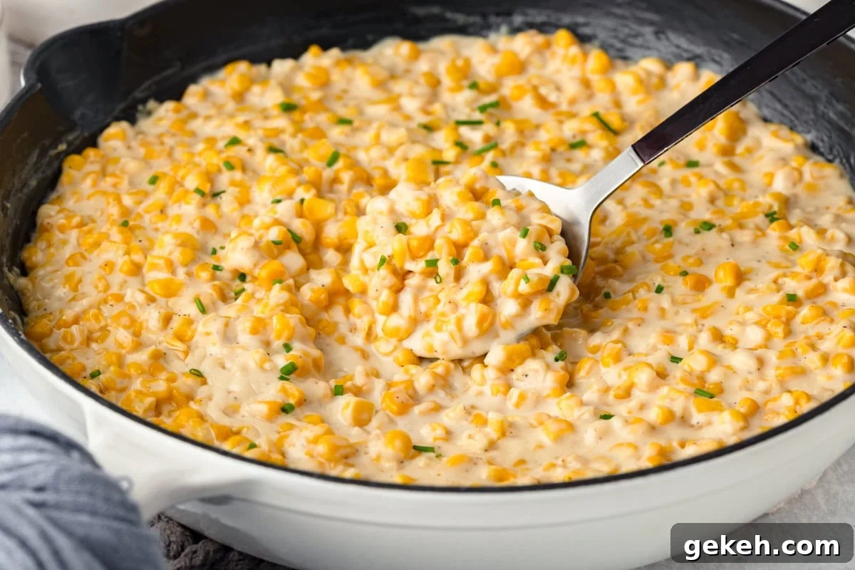 A white skillet filled with creamy corn decorated with small green chives. A spoon is lifting a portion, showing the rich, thick texture of the dish. The skillet rests on a light surface with a gray cloth partially visible.