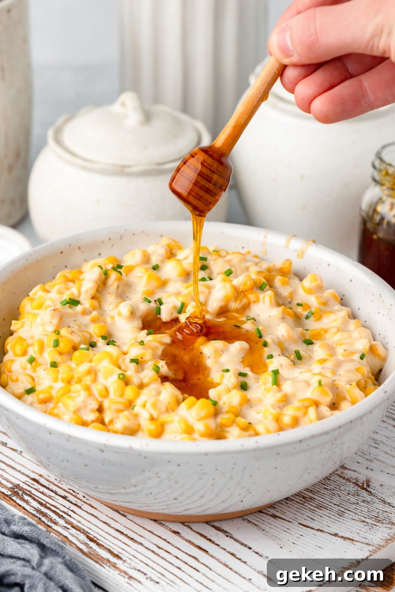 A hand drizzles honey over a bowl of creamy corn salad topped with chopped chives. The bowl is set on a wooden surface with ceramic jars in the background.