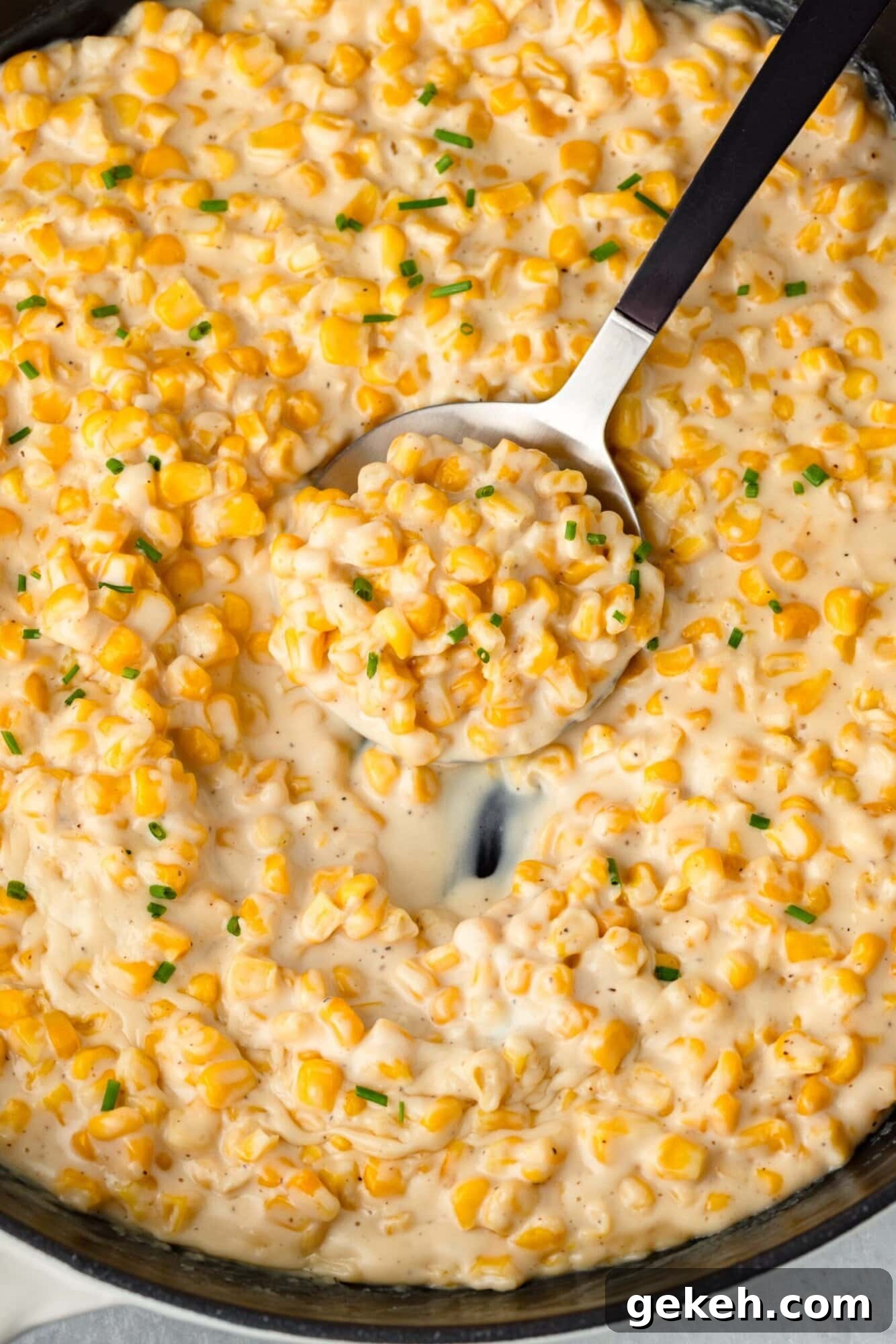 A skillet filled with creamy corn mixture, garnished with chopped chives. A spoon is lifting a portion, showcasing the creamy texture and vibrant yellow corn kernels.