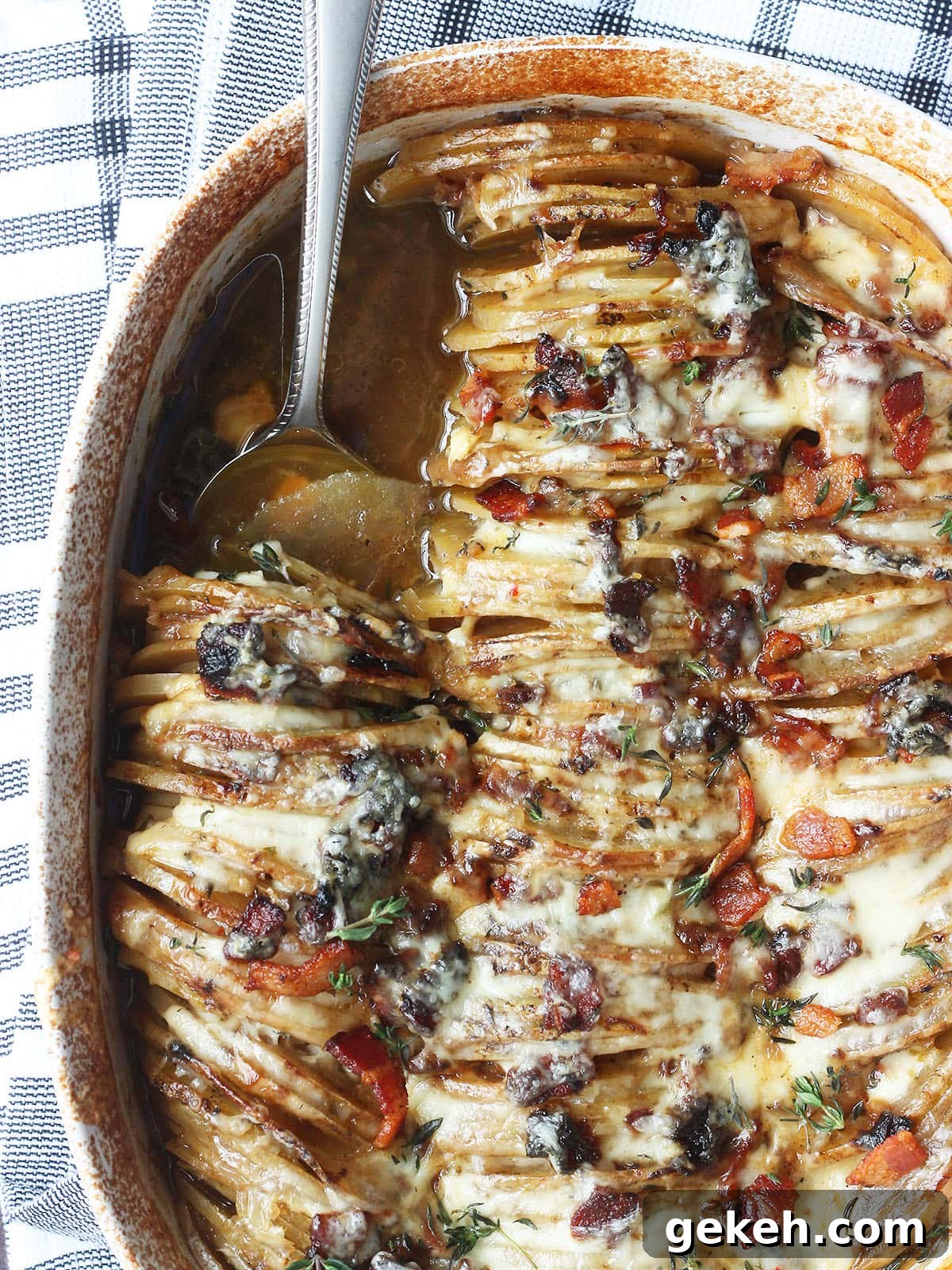 Creamy Scalloped Potatoes with Crispy Bacon 25 Overhead shot of a metal spoon scooping out a serving of scalloped potatoes with bacon from a white oval casserole dish.