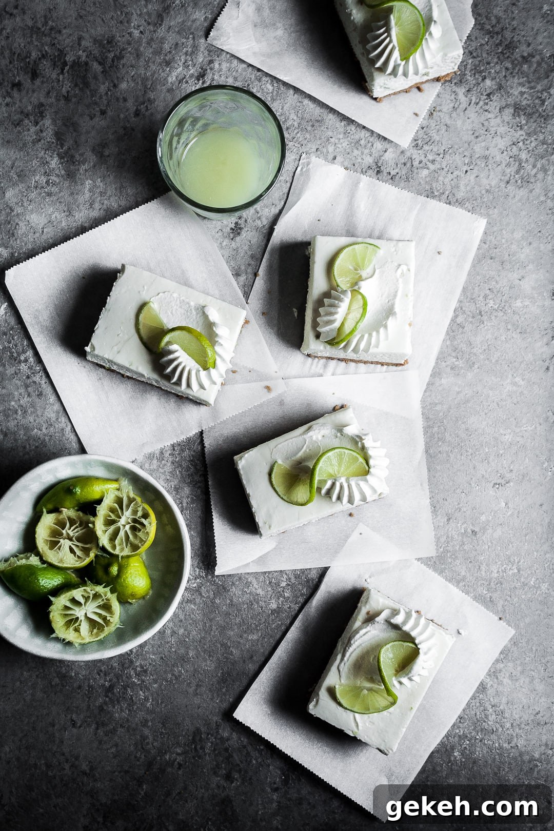 No-Bake Key Lime Cheesecake Bars Recipe for Zesty Dessert 2 Cheesecake bar slices with piped whipped cream and lime twists on squares of parchment paper on a grey surface.