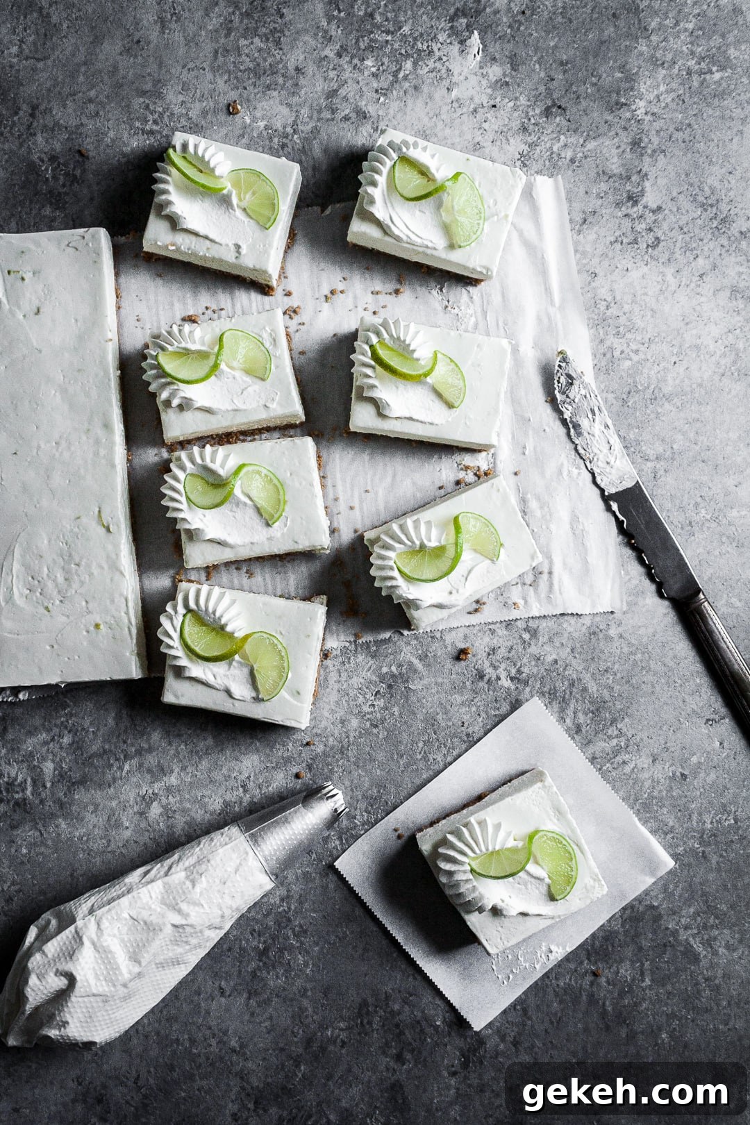 No-Bake Key Lime Cheesecake Bars Recipe for Zesty Dessert 11 Five sliced cheesecake bars on parchment paper squares on a grey background with a piping bag of whipped cream and vintage knife nearby