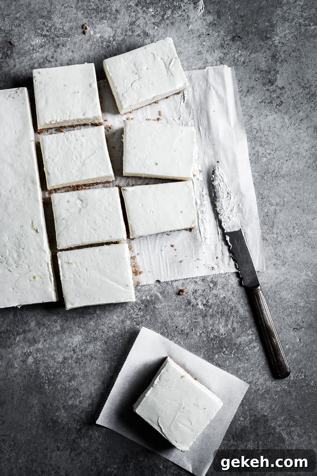 No-Bake Key Lime Cheesecake Bars Recipe for Zesty Dessert 7 Sliced cheesecake bars on parchment paper squares on a grey background with a vintage knife nearby