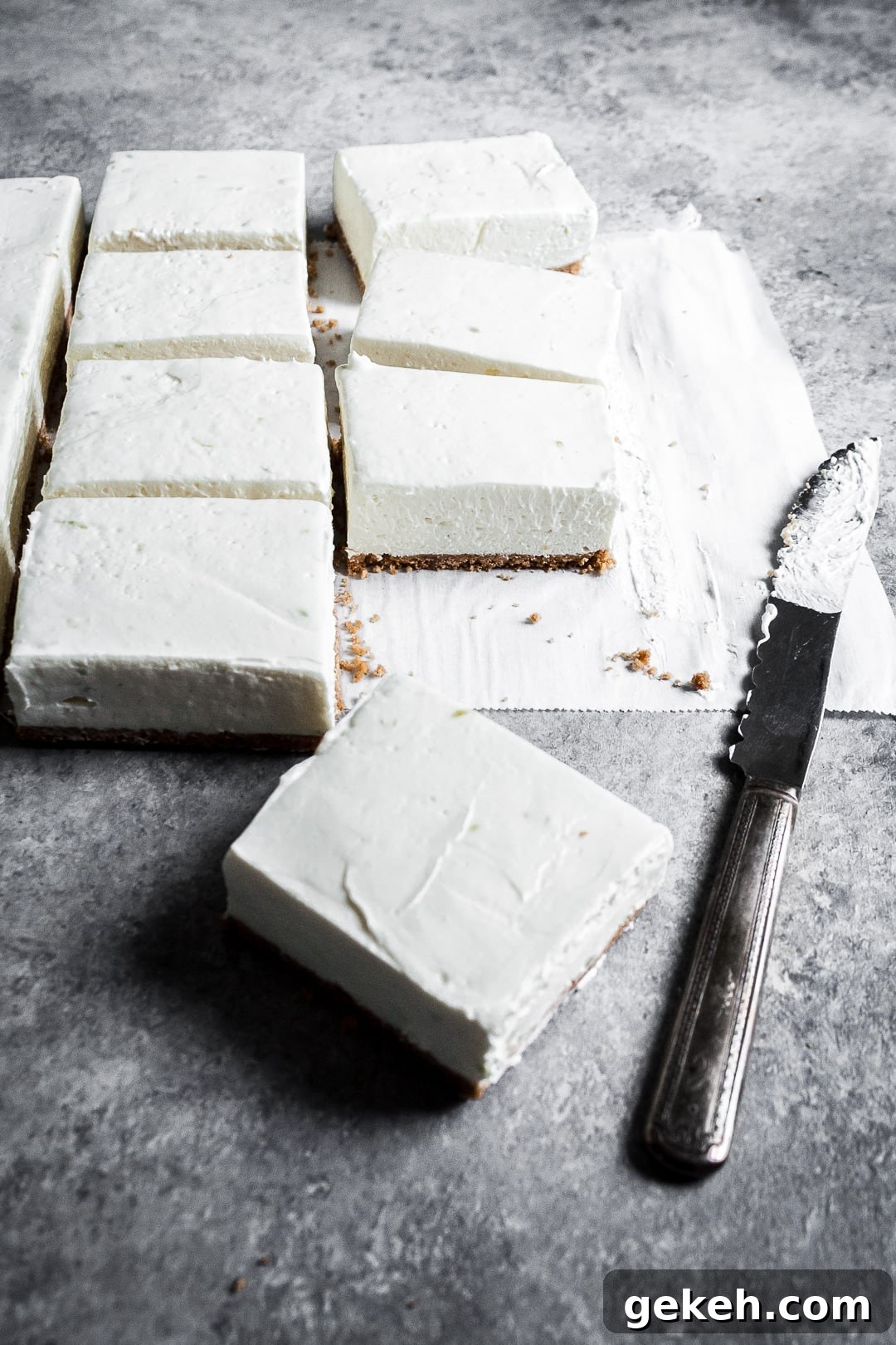 No-Bake Key Lime Cheesecake Bars Recipe for Zesty Dessert 8 45 degree angle view of sliced cheesecake bars on parchment paper squares on a grey background with a vintage knife nearby
